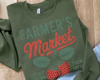 May include: Green sweatshirt with the text "FARMER'S Market" in red and green lettering. The text is distressed and has a vintage look. There are two red strawberries with green leaves on the bottom of the sweatshirt. The text "LOCALLY GROWN" and "FARM FRESH" are also on the sweatshirt.