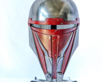 May include: A red and silver Mandalorian helmet with a weathered finish. The helmet is on a black stand with a gear design.
