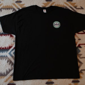 May include: Black crew neck t-shirt with a small circular logo on the upper left chest. The logo features the word "Heineken" in white, green, and red, with text around the logo. The shirt is made of a soft material.