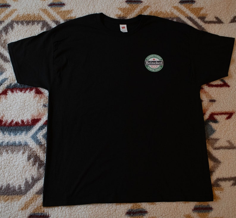 May include: Black crew neck t-shirt with a small circular logo on the upper left chest. The logo features the word "Heineken" in white, green, and red, with text around the logo. The shirt is made of a soft material.