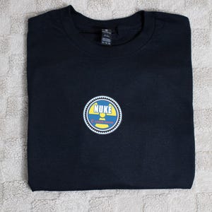 Könnte beinhalten: Ein marineblaues T-Shirt mit einem kreisförmigen Aufdruck auf der Vorderseite. Der Aufdruck zeigt das Wort "NUKE" in Gelb, Blau und Weiß mit einem Strahlungssymbol. Das T-Shirt ist sauber auf einer hellen Oberfläche gefaltet.