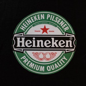 May include: A circular Heineken Pilsener logo on a black backdrop. The logo has a green border with white text and a red star. The text includes "Heineken", "Premium Quality", and "Trade Mark".