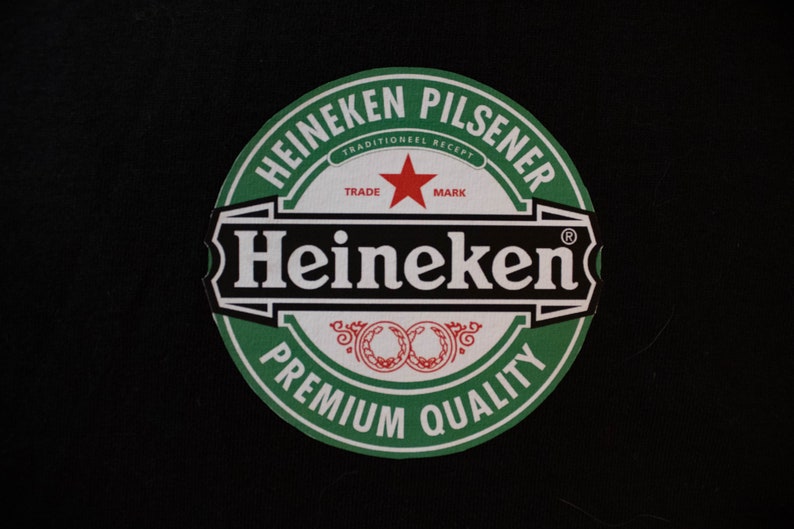 May include: A circular Heineken Pilsener logo on a black backdrop. The logo has a green border with white text and a red star. The text includes "Heineken", "Premium Quality", and "Trade Mark".