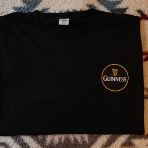 Puede incluir: Camiseta negra lisa con un peque&ntilde;o logotipo circular de Guinness en el pecho derecho. El logotipo presenta un arpa dorada y la palabra "Guinness" en blanco, sobre un fondo negro y dorado. La camiseta tiene cuello redondo.