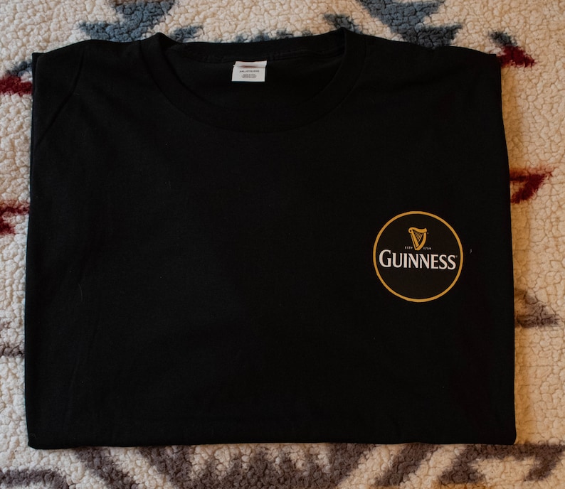Puede incluir: Camiseta negra lisa con un peque&ntilde;o logotipo circular de Guinness en el pecho derecho. El logotipo presenta un arpa dorada y la palabra "Guinness" en blanco, sobre un fondo negro y dorado. La camiseta tiene cuello redondo.