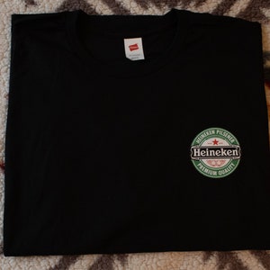 May include: Black t-shirt featuring the Heineken Pilsener logo, a green circular design with white text and a red star. The shirt is made of a soft material and has a tag.