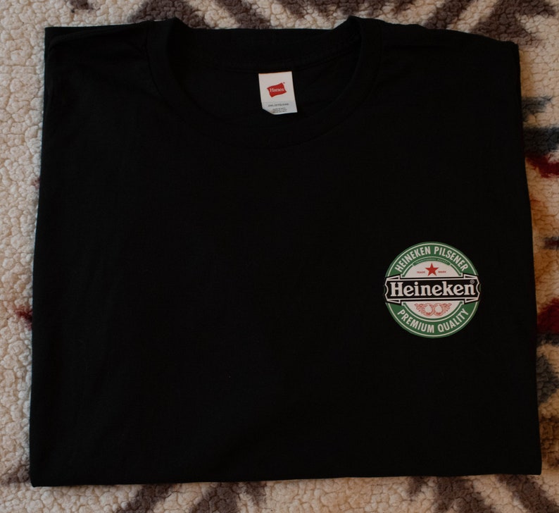 May include: Black t-shirt featuring the Heineken Pilsener logo, a green circular design with white text and a red star. The shirt is made of a soft material and has a tag.