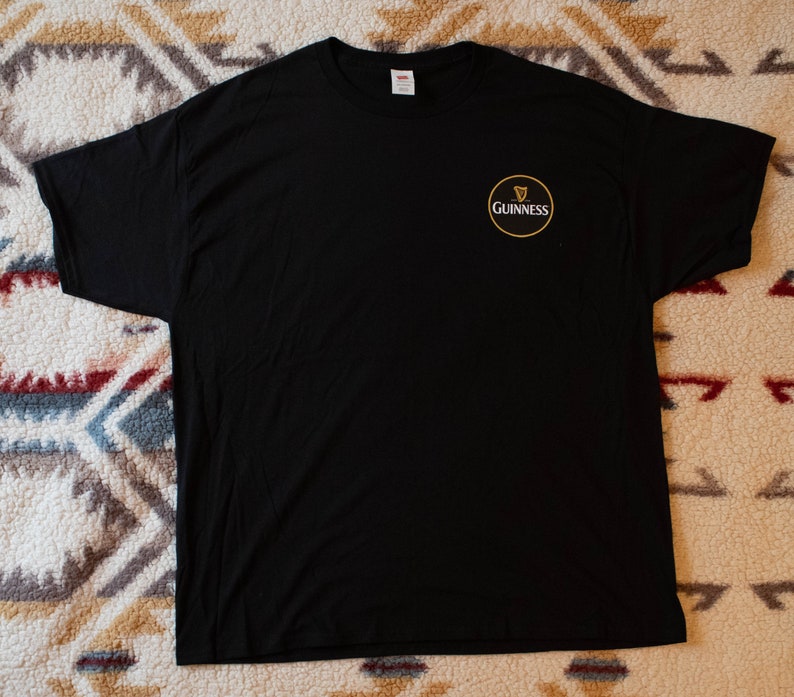 Puede incluir: Camiseta negra con el logo de Guinness en el pecho izquierdo. El logo es un c&iacute;rculo dorado con el s&iacute;mbolo del arpa Guinness y la palabra "Guinness" en blanco. La camiseta es de tejido suave.