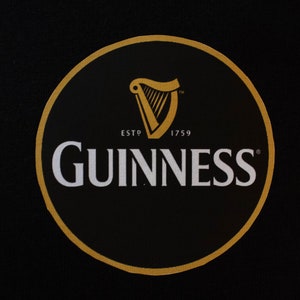 Puede incluir: C&iacute;rculo negro con borde dorado que muestra el logotipo de Guinness. El logotipo incluye un arpa dorada, la palabra "GUINNESS" en blanco y el texto "ESTD 1759".