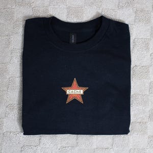 Könnte beinhalten: Gefaltetes schwarzes T-Shirt mit einem roten und goldenen Stern-Design. Der Stern hat das Wort "CACHE" in einem weißen Rechteck aufgedruckt. Das Shirt besteht aus einem weichen Material.