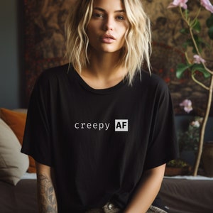 May include: A woman with blonde hair is wearing a black t-shirt with the text "creepy AF" printed on it in white.
