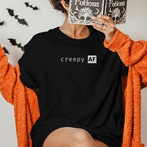May include: A black long-sleeved t-shirt with the text "creepy AF" printed in white on the front. The person is wearing a bright orange fuzzy cardigan and black and orange socks.