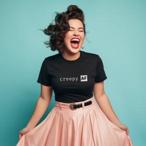 May include: A woman wearing a black t-shirt with the text "creepy AF" in white letters. She is wearing a pink skirt and has her hair styled in loose curls. She is laughing with her mouth open.