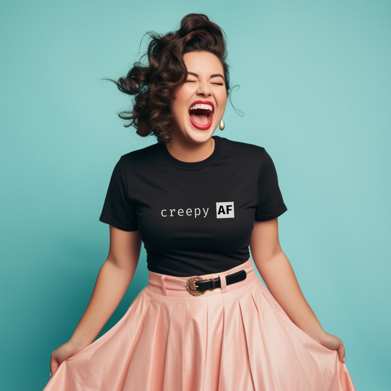 May include: A woman wearing a black t-shirt with the text "creepy AF" in white letters. She is wearing a pink skirt and has her hair styled in loose curls. She is laughing with her mouth open.