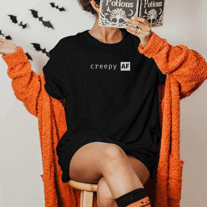 May include: A black long-sleeved t-shirt with the text "creepy AF" printed in white on the front. The person is wearing a bright orange fuzzy cardigan and black and orange socks.