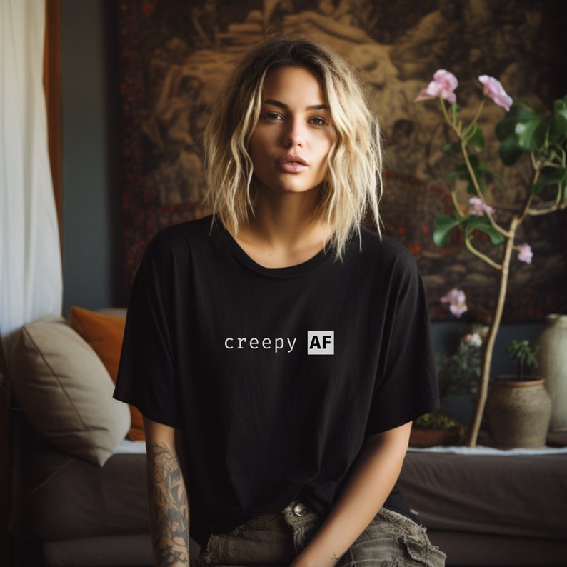 May include: A woman with blonde hair is wearing a black t-shirt with the text "creepy AF" printed on it in white.