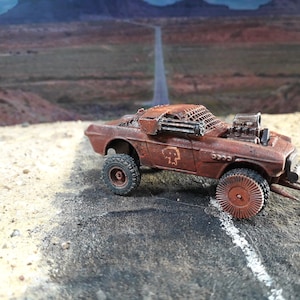 Puede incluir: Un coche de juguete marrón oxidado, de estilo post-apocalíptico, con neumáticos grandes y un emblema de calavera. El coche tiene un motor modificado y una estructura en forma de jaula en la parte superior. Está aparcado en una carretera agrietada con un paisaje desértico al fondo.