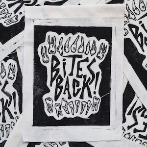 May include: A black and white square patch with the words "BITES BACK!" in a bold, stylized font. The design features illustrations of teeth around the text. The patch is on a white fabric background, with other similar patches visible.
