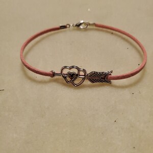 Puede incluir: Un brazalete de gamuza rosa con un dije de plata con forma de corazón doble con una flecha que lo atraviesa.