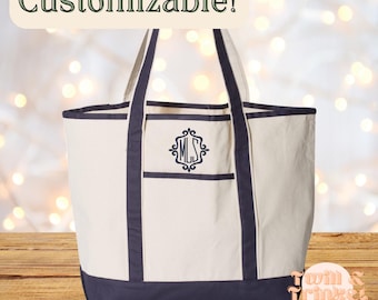 May include: Cream-colored canvas tote bag with navy blue accents. Features a front pocket with a monogram and the text "Customizable!". The bag has navy blue handles and trim. The bag is sitting on a wooden surface.