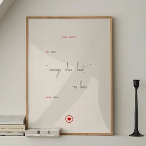 May include: A framed print with the words "courage, dear heart" in elegant script. The print is in a light wood frame and has a minimalist design. Two black candle holders and a stack of books are on the shelf below.