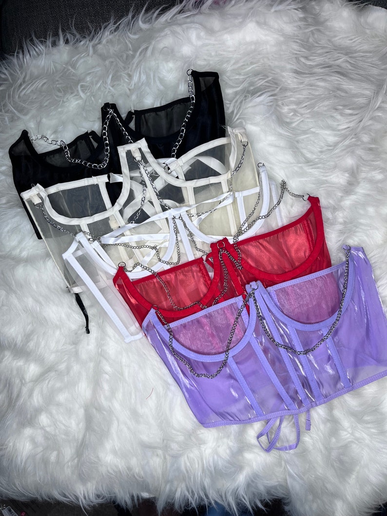 Op de afbeelding: Vijf doorschijnende mesh tops met kettingbandjes in verschillende kleuren: zwart, wit, cr&egrave;me, rood en paars. De tops zijn op elkaar gelegd, met de zwarte top onderaan en de paarse top bovenaan.
