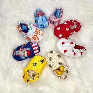Puede incluir: Una colección de zapatos de bebé en varios patrones y colores. Los diseños incluyen corazones rojos, círculos azules, flores rosas, coral blanco, náutico azul marino, abejas amarillas y panales blancos. Los zapatos están colocados sobre una superficie blanca y esponjosa.