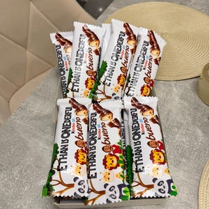 Könnte beinhalten: Eine Schachtel Kinder Bueno Schokoriegel. Die weißen Verpackungen tragen den Text "ETHAN IS ONEDERFUL" und "Kinder bueno" mit Cartoon-Tierillustrationen. Die Riegel sind in einer rechteckigen Schachtel angeordnet.