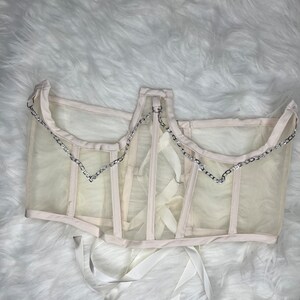 Op de afbeelding: Een wit mesh-corset met een zilveren kettingdetail en witte lintjes.