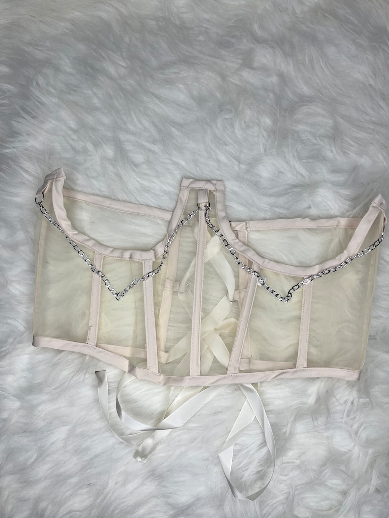 Op de afbeelding: Een wit mesh-corset met een zilveren kettingdetail en witte lintjes.