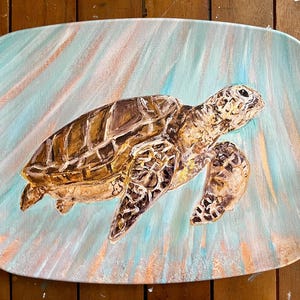 May include: An oblong, hand-painted canvas features a sea turtle swimming in a turquoise and peach-colored ocean. The turtle has a brown shell with a patterned design and a light-colored head and flippers. The artwork is a decorative piece.
