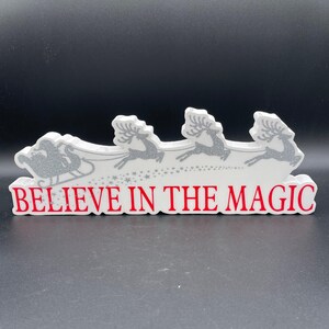 May include: A white Christmas decoration featuring Santa's sleigh pulled by reindeer. The sleigh and reindeer have a silver glitter design. The words "BELIEVE IN THE MAGIC" are in red. The decoration is on a black surface.