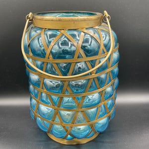 May include: A decorative blue glass lantern with a brass handle and accents. The lantern features a woven brass cage design around the blue glass body. The handle is arched, allowing for easy carrying or hanging. The lantern is designed for indoor or outdoor use.