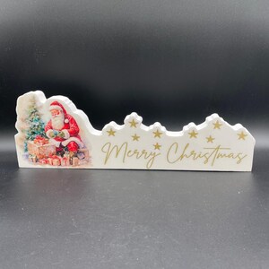 May include: A white Christmas decoration with a Santa Claus illustration and the text "Merry Christmas" in gold script. The decoration features a depiction of Santa Claus with gifts and a Christmas tree. Gold stars are also included.
