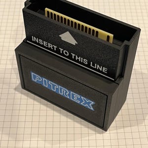May include: Black plastic Pitrex cartridge with a cream-coloured connector. The cartridge has the text "INSERT TO THIS LINE" and the Pitrex logo in blue. The cartridge is designed to hold a game or program.