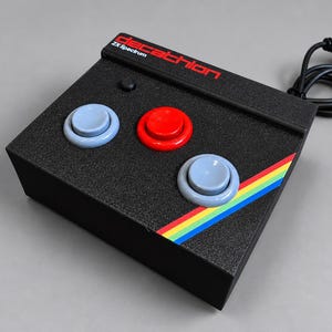 Puede incluir: Un mando de videojuegos rectangular negro con el texto Decathlon ZX Spectrum en rojo y blanco. El dispositivo tiene dos botones azul claro, un botón rojo central, un botón negro pequeño y un patrón de rayas diagonales de arcoíris.