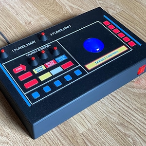 May include: A black retro-style game controller with a blue trackball and various buttons labelled "Fire," "Alpha Base," "Delta Base," and "Omega Base." It has a "Target Control" section and a "Launch Control" area. The controller is on a wooden surface.