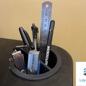 May include: A black desk organiser with a round compartment for pens and pencils, and a rectangular compartment for a ruler. The organiser contains a black stylus, a ruler marked in centimetres, and two silver USB drives with the Stratasys logo.