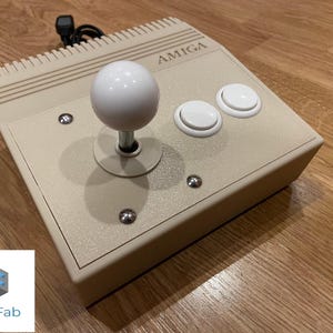 Könnte beinhalten: Beige AMIGA-Joystick-Controller mit einem weißen, kugelförmigen Stick und zwei weißen Knöpfen. Der Controller hat eine strukturierte Oberfläche und ist mit silbernen Schrauben befestigt. Ein schwarzes Kabel erstreckt sich von der Rückseite.