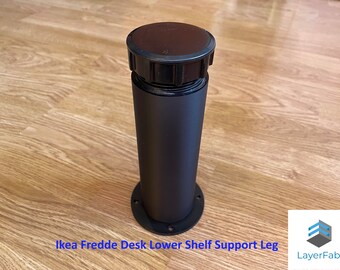 May include: A black Ikea Fredde desk lower shelf support leg. The cylindrical leg has a black base with screw holes and a black top. The leg is made of metal and is designed to support a desk shelf.