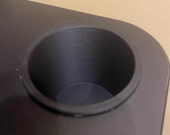 May include: Close-up of a black, built-in cup holder. The cup holder is circular and recessed into a black surface. The interior of the cup holder has a textured design. The background is a light beige wall.