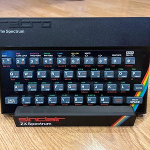 Puede incluir: Un ordenador Sinclair ZX Spectrum negro con un teclado completo y detalles de arcoíris. El teclado tiene varias teclas de función y un diseño retro, con el texto "The Spectrum" en la parte superior.