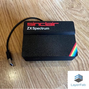 Puede incluir: Dispositivo negro Sinclair ZX Spectrum con texto rojo y una franja de arcoíris. El dispositivo tiene un cable USB. El dispositivo está sobre una superficie de madera.