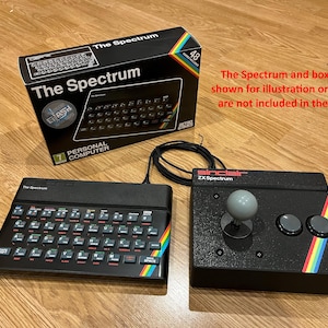 Puede incluir: Ordenador Sinclair ZX Spectrum negro con teclado y joystick. El teclado tiene teclas de colores y una franja arcoíris. El joystick es negro con una bola y botones grises. También se muestra una caja negra con "The Spectrum".