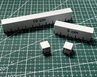 May include: Four white plastic rulers, two are 1 cm cubes, one is 5 cm long, and one is 10 cm long. The rulers are on a green cutting mat.