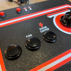 May include: Black arcade game controller with red and white accents. Features a large black knob, several black buttons, and red buttons. The text "FIRE" and "SUPER ZAPPER" are visible.