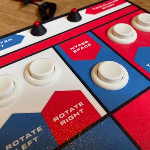 Puede incluir: Primer plano de un mando de juego arcade retro con botones blancos y detalles en rojo y azul. El mando tiene etiquetas como "ONE PLAYER START", "HYPER SPACE" y "THRUST".