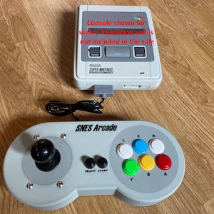 Pode incluir: Uma consola Super Nintendo e um comando SNES Arcade são exibidos numa superfície de madeira. O comando tem um joystick e botões coloridos. A consola é mostrada para escala e não está incluída na venda.