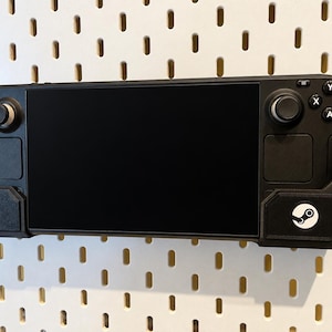 May include: A black handheld gaming console secured in a black wall-mounted holder. The device features a large rectangular screen, a directional pad, two analog sticks, four action buttons labelled A, B, X, and Y, and two trackpads. A blue and white logo is visible.