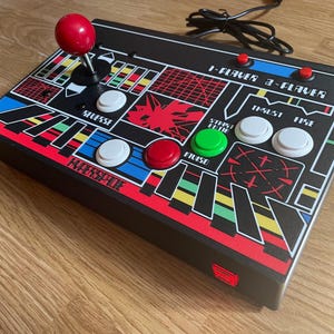 May include: Black arcade game controller with a red joystick and buttons. The controller has a colourful graphic design with the text "HYPERSPACE" and other game-related labels. It features buttons for 1 and 2 players.